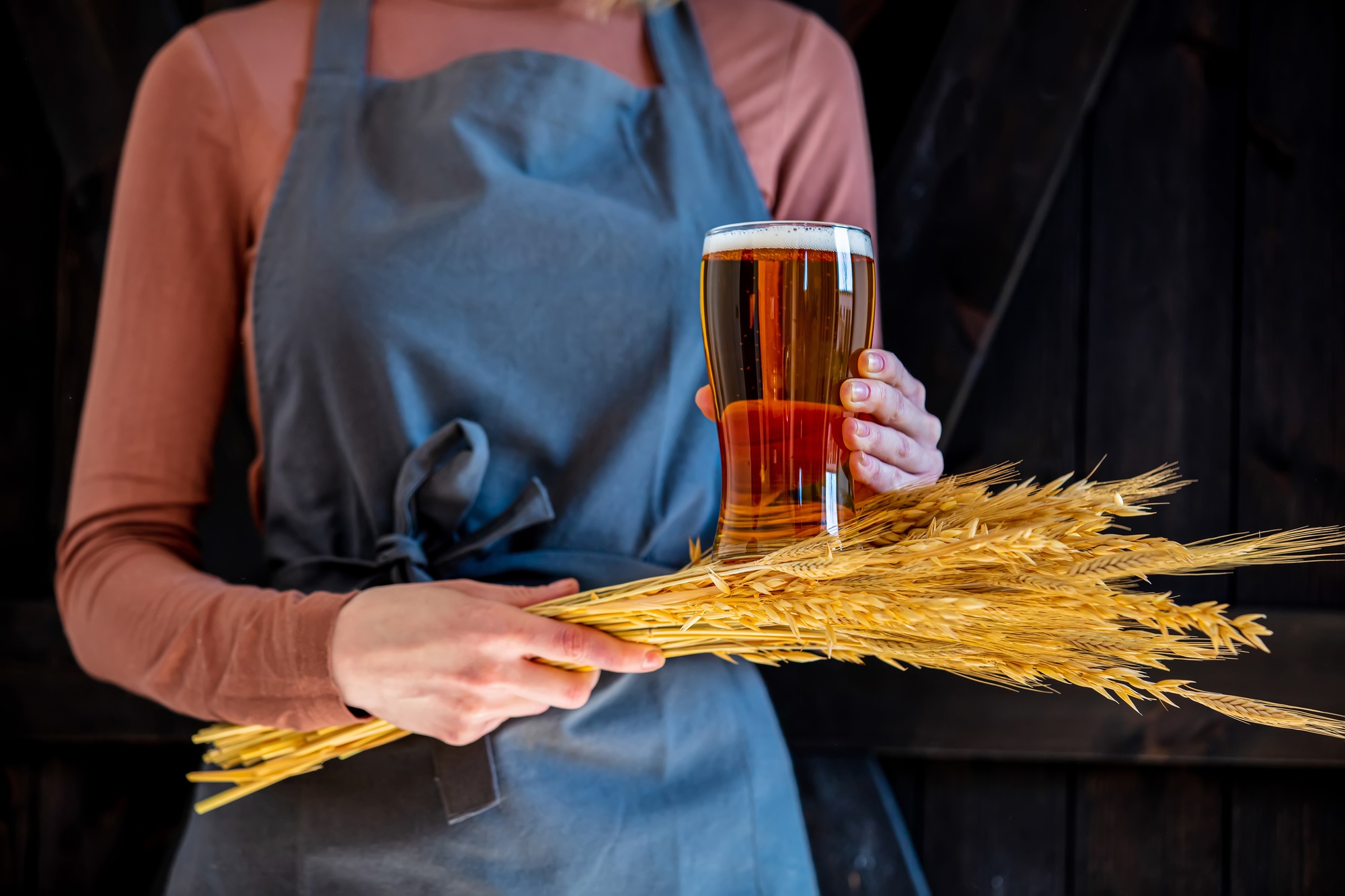 brewer in apron holds glass of beer and wheat spikelets